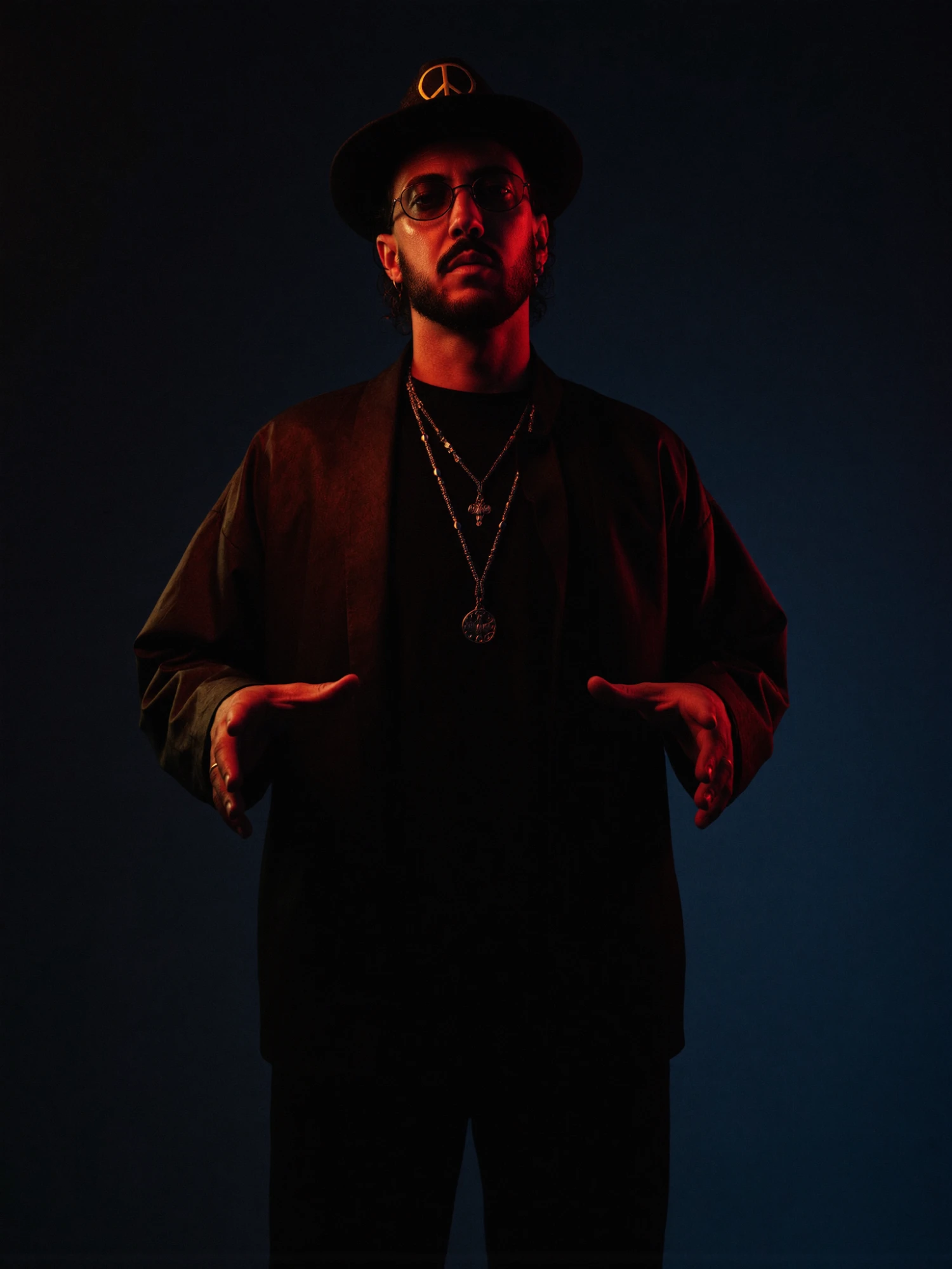 Fudge Jarcheh in a wide-brim peace-sign hat and round glasses, hands open in front of him, lit with a warm amber rim on one side and a cool deep-blue ambient behind.